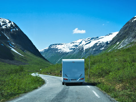 Camper RV Trailer Caravan On Asphalt Road In Norway, Back Side, In Mountains With Snow Green Trees Blue Sky And Clouds.