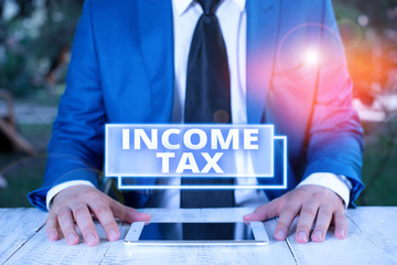 Writing note showing Income Tax. Business concept for Annual charge levied on both earned and unearned income Businessman in blue suite with a tie holds lap top in hands