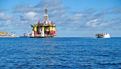 A huge offshore oil rig enters a harbour, a fast ferry leaves the harbour, various service ships at work, blue sea and sky over a commercial port.