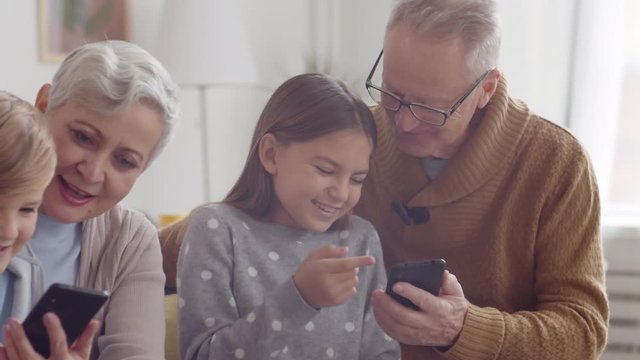 Close up of Caucasian aged family couple sitting on couch in living room with their grandkids and using smartphones