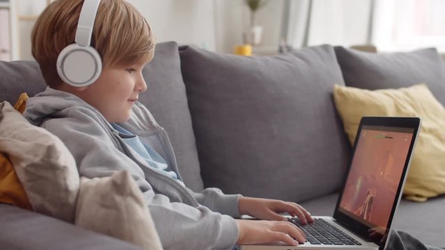 Medium shot of Caucasian boy wearing grey hoody and headphones sitting on sofa with laptop on his knees and playing computer game
