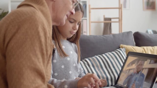 Close up of teenage Caucasian girl sitting with her grandfather on sofa in living room and showing him different videos on PC