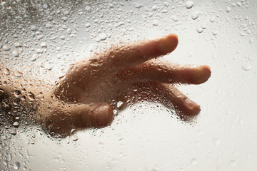 hand of a young woman behind glass with water drops on it reaching out