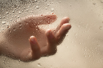 hand of a young woman behind glass with water drops on it reaching out