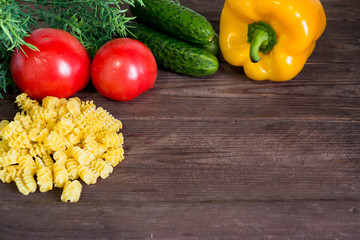 Macaroni, tomatoes, cucumber, dill and bell pepper on a dark wooden background. Ingredients for cooking. Look at the bench.