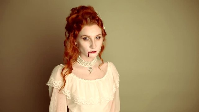 Young vampire woman with long curly hair, pale skin in a white dress on a black background. A beautiful redhead model with red lips. Outfit for halloween. Vampire with open mouth and fangs