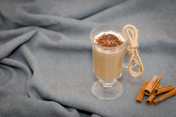 Traditional winter eggnog in glass mug with milk, cinnamon and chocolate covered with whipped cream on a gray cloth,cinnamon sticks on cloth background. Advertising for cafe menu. Coffee.