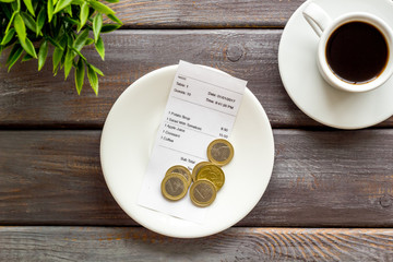 Pay restaurant bill by cash. Reciept and coins on plate on dark wooden background top view