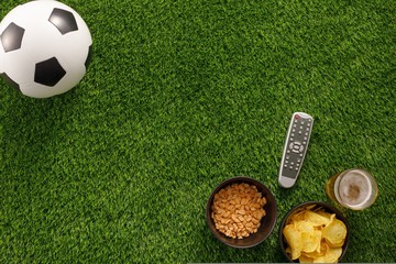 Naklejka premium Soccer ball on a green field and ottoman for a fan with snacks and a TV remote control. flat lay.Copy space.