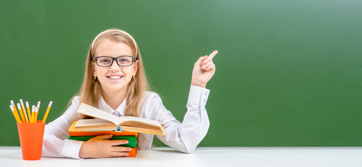 Smiling young girl wearing a eyeglasses hugs books and points away on empty green chalkboard. Empty space for text - Powered by Adobe