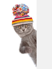 Kitten wearing a warm hat with pompon peeks behind empty banner. isolated on white background