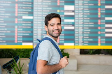 Cheerful bearded man standing near departure board. Smiling Hispanic traveler looking at camera. Travel concept