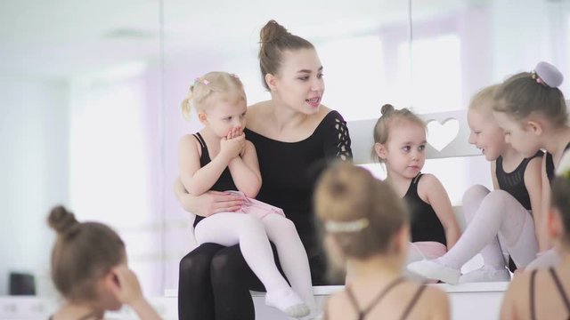 Young ballet teacher sitting on bench in ballet school with little girls in black leotards and communicating with them