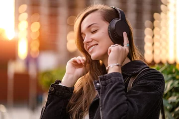 Fotobehang Happy joyful stylish casual fashionable hipster student woman teenager enjoys listening music using black wireless headphones while walking around the city. Music lover © Goffkein