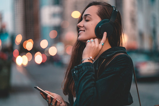 Young happy stylish trendy casual hipster woman teenager listening to music on a black wireless headphone while walking around the city. Music lover enjoying music