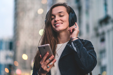 Young happy stylish trendy casual hipster woman changes songs and tracks on smartphone during listening to music on a wireless headphone while walking around the city. Music lover enjoying music