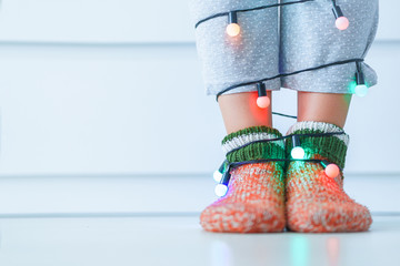Female legs in warm knitted soft cozy christmas socks and garland with illuminated lights in wintertime at home. Copy space