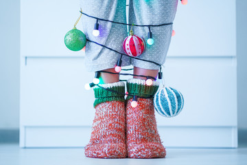 Female legs in warm knitted soft cozy christmas socks, baubles balls and garland with illuminated lights in wintertime at home.