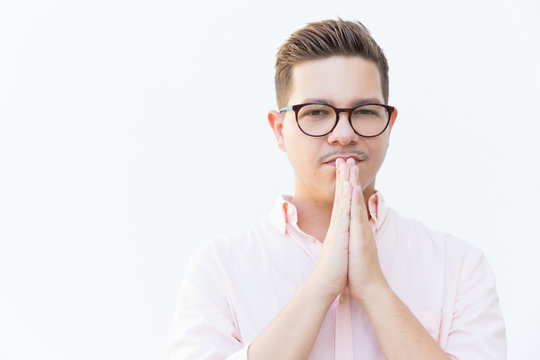 Serious pensive guy in eyewear keeping hands at face together and looking at camera. Young man in glasses standing isolated over white background. Pray gesture concept