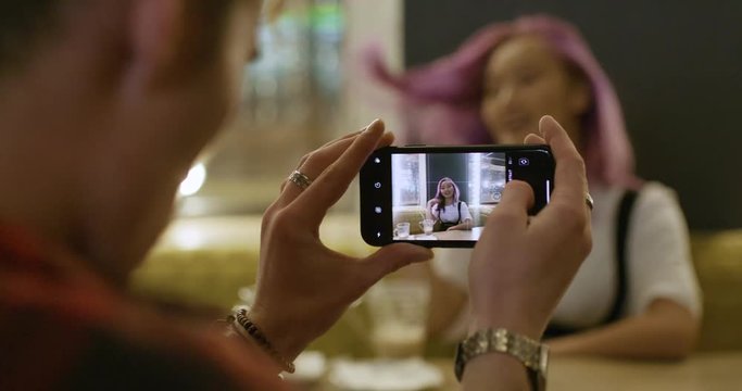 Young couple in a modern caffe, man photographs a asian girl with pink hair on the smartphone, teenagers have a rest in the city.