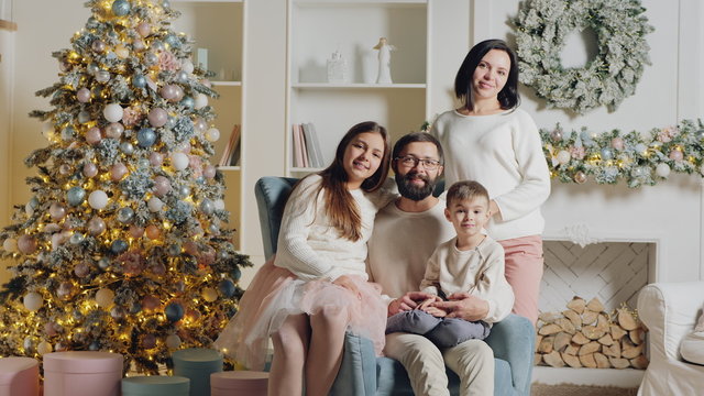 Cute European family of four posing on camera for a Christmas photo in the living room of his house in an armchair near the Christmas tree and fireplace