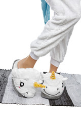 Close-up shot of female legs in white velour trousers and plush house slippers made in the form of white smiling unicorn. The girl is standing on the striped gray and white carpet.