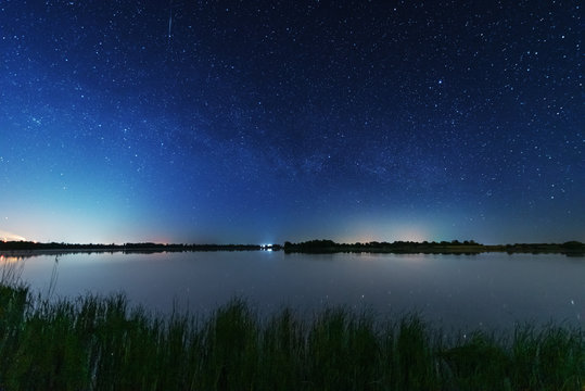 A magical starry night on the river bank with a large tree and a milky way in the sky and falling stars in the summer.