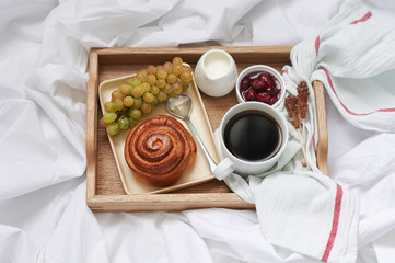 Breakfast on white bed sheets in hotel room. Close up of wooden tray with coffee, bun, jam and grape
