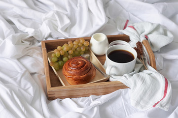 Breakfast on white bed sheets in hotel room. Close up of wooden tray with coffee, bun, jam and grape