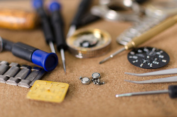 Closeup of tweezers and little watch batteries, clock faces, screwdrivers, pieces of dial,  wrist watches on the brown surface