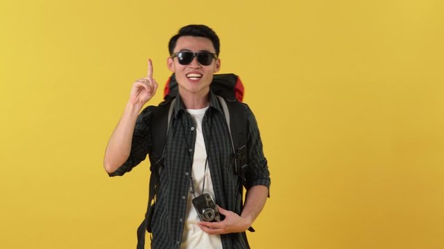 Young happy smiling Asian man with backpack and sunglasses taking photos and waving hand against yellow background