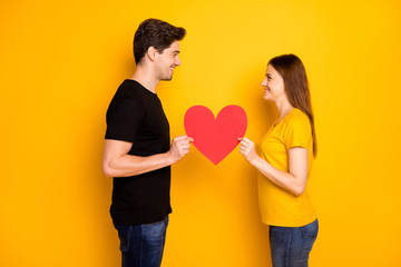 Fototapeta na wymiar Photo side profile of two beloved cheerful charming cute nice couple wearing jeans denim black t-shirt looking into each other eyes holding red heart isolated vivid color background