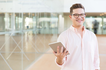 Joyful tablet user in glasses happy about high speed connection in public space. Young man with tablet posing indoors with glass wall interior in background. Internet concept