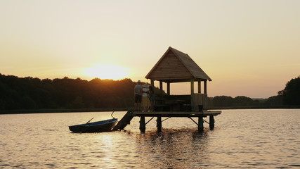 A family of three stand near a wooden gazebo in the middle of the lake, hugging together watching the sunset