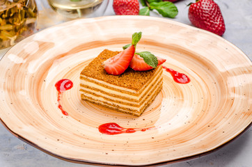 Delicious piece of honey cake served with fresh summer berry on a plate, close up on a background teapot. Homemade russian Medovik decorated with mint and strawberry.