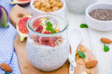 Chia pudding with yogurt, homemade granola, fresh figs and honey in a glass jar on a board on a white wooden background.