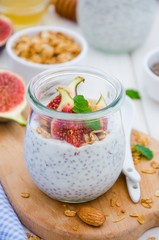 Chia pudding with yogurt, homemade granola, fresh figs and honey in a glass jar on a board on a white wooden background.