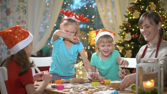 Woman and three little girls baking Christmas cookies. Children with their mom making gingerbread for family dinner on Christmas eve in dining room with christmas tree and candles.
