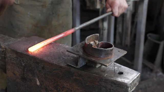 hands of a male blacksmith takes from a furnace red-hot iron with hammer blows forms on a large anvil in a blacksmith workshop