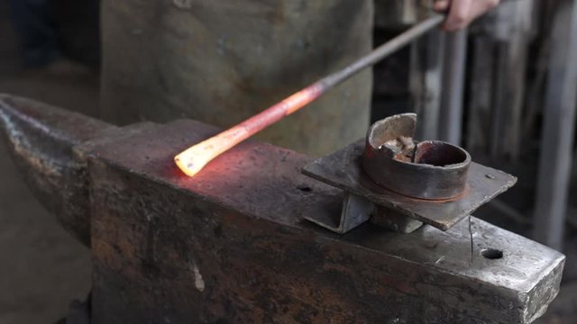 hands of a male blacksmith takes from a furnace red-hot iron with hammer blows forms on a large anvil in a blacksmith workshop