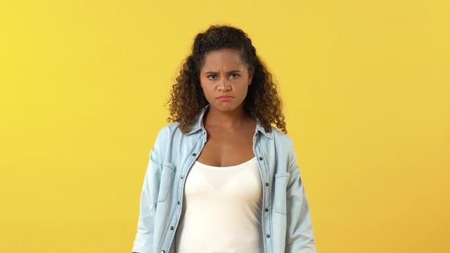 Young unhappy African American woman acting unsure and displeased while shrugging shoulders and folding arms against yellow background