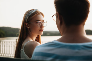 Charming young woman with red hair and freckles talking with her boyfriend outdoor while sitting on a beach in the park.