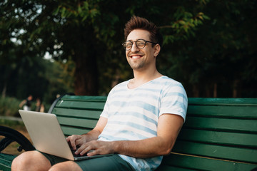 Portrait of a young man looking at camera smiling while holding a laptop on his legs outside in the park.