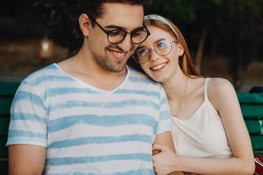 Portrait of a beautiful couple embracing while sitting on a bench outdoor while woman with red hair and freckles is looking at hims smiling leaning her head.