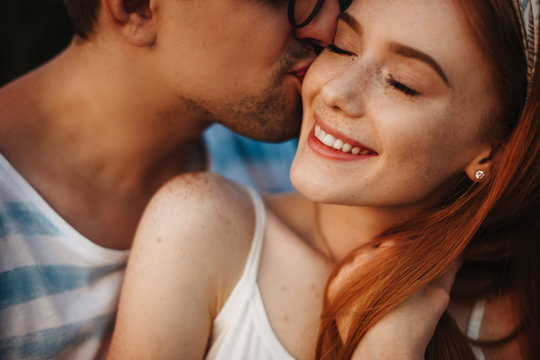 Close up of a lovely young woman with red hair an frekles laughing while eyes closed while is kissed by her boyfriend outdoor while dating.