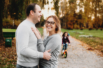 Portrait of a lovely father and mother embracing and laughing while thier daughter is riding a bike in the park.
