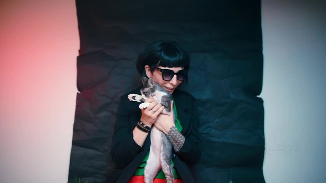 A girl with black hair and black glasses holds a Devon Rex cat in her arms.