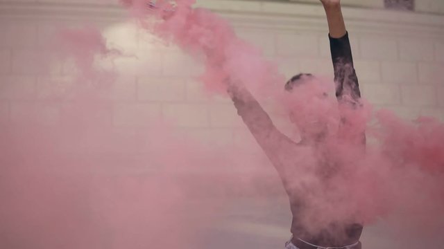 Beautiful afro american woman with pink smoke bomb or grenade on bright bricked wall background. Girl in black sweater and jeans waving grenade with a hand and relaxfully dancing. Slow motion