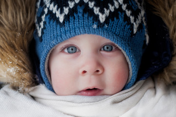 face of a beautiful baby with blue eyes close-up, in a blue knitted hat, in a white scarf and a hood with fur, frozen pink cheeks in winter