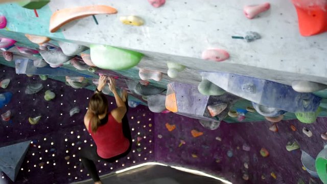 Wide shot of young athletic Caucasian woman in sports top and leggings bouldering on advanced route of artificial indoor climbing wall, hanging on hands off overhang then pulling herself up
